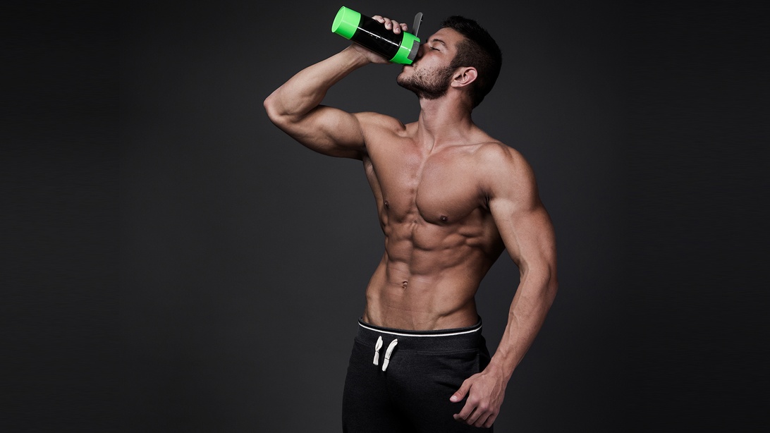 En muskulös individ med bar överkropp dricker ur en svart och grön proteinshaker mot en mörk bakgrund.